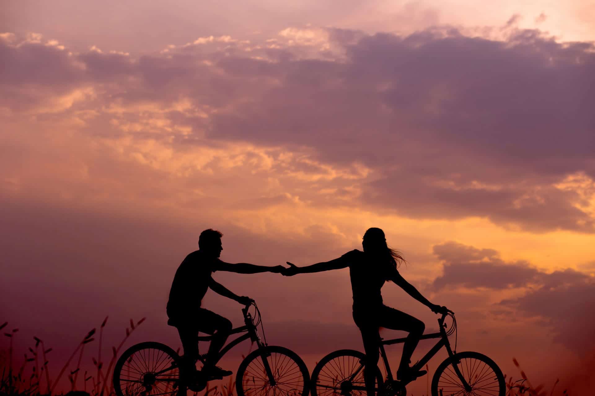 Par som cyklar i solnedgång och sträcker sina händer mot varandra